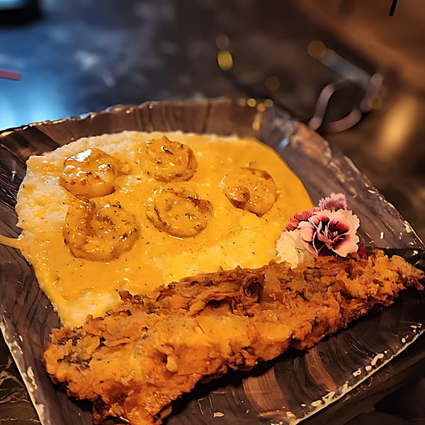 Southern shrimp and grits with fried fish and a pink flower garnish on a dark plate.