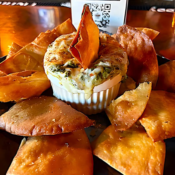 A white ramekin of cheesy spinach dip with a chip, surrounded by golden fried pita bread.