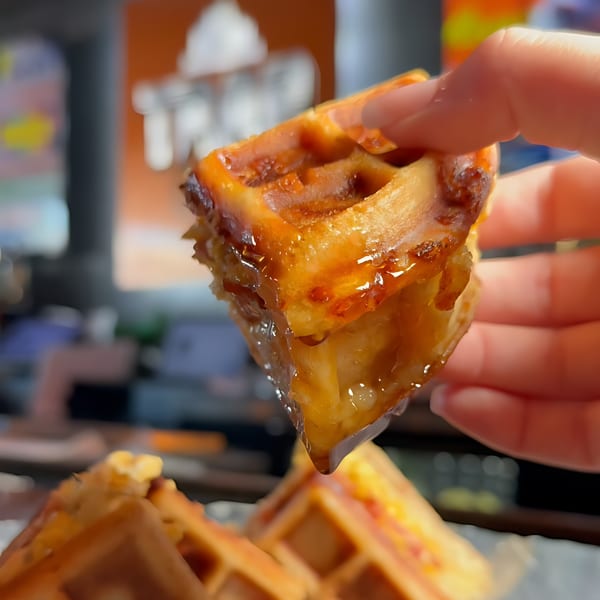 A hand holds a golden-brown waffle bite, dripping with sweet syrup, at Trap City Cafe.